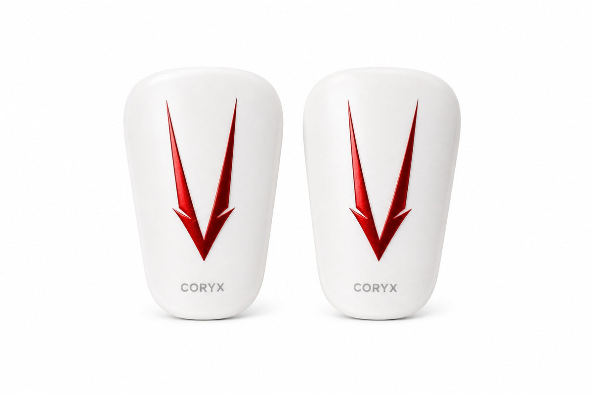 Two white CORYX shin pads shown side by side on a clean white background, each featuring a large red chevron logo and the CORYX brand name near the bottom.